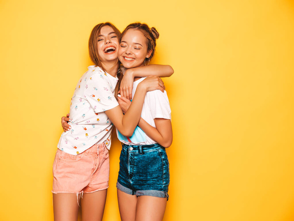 Two young beautiful smiling hipster girls in trendy summer clothes. Sexy carefree women posing near yellow wall in studio. Positive models going crazy and having fun.Hugging - Zdjęcie, obraz