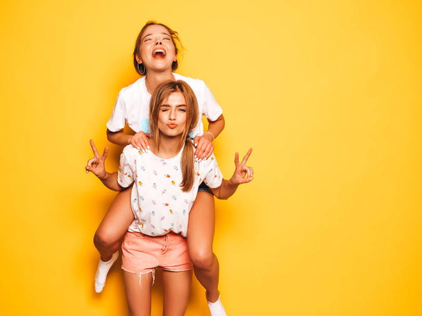 Two young beautiful smiling hipster girls in trendy summer clothes. Sexy carefree women posing near yellow wall in studio.Model sitting on her friend's back and shows peace sign - Foto, Bild