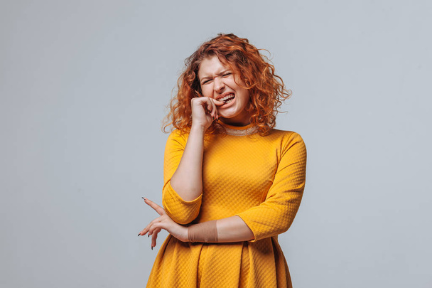 Red-haired girl bites her nails on a light gray background - Fotó, kép