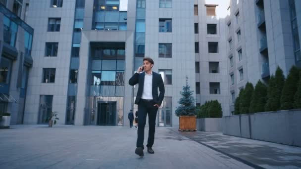 erfolgreicher Geschäftsmann, der auf den Straßen des Geschäftsviertels spaziert und mit dem Handy telefoniert. selbstbewusste attraktive kaukasische Mann im Anzug zu Fuß nach draußen in der Nähe von großen Bürogebäuden und mit Smartphone. - Filmmaterial, Video