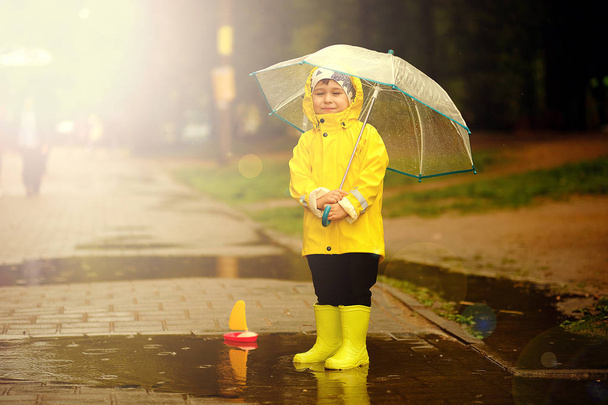 ευτυχισμένο παιδί παίζει σε μια λακκούβα σε μια βροχερή καλοκαιρινή μέρα. ένα αγόρι με ένα κίτρινο αδιάβροχο περπατάει στο πάρκο.. - Φωτογραφία, εικόνα