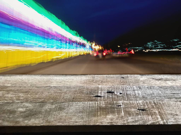 Perspektywny drewniany blat i rozmyty kolorowy bokeh z oświetleniem ulicznym w nocy, kropla deszczu na przedniej szybie samochodu.Jazda samochodem w ulewny deszcz.Ruch w mieście w deszczowy dzień, wolne miejsce na tekst - Zdjęcie, obraz