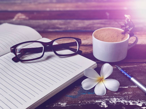 Eyeglasses on book and White mugs has Cappuccino coffee on old wood,rustic still life,soft dark tone,dimly light,with lens flare,free space for your text design. Concept of Relaxation in Coffee time. - Photo, Image