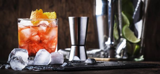 Red cocktail with dry vermouth, bitter, soda, orange zest and ice, wooden bar counter background, bar tools, selective focus - Фото, изображение