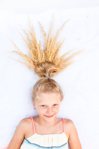 niedlich lächelndes kleines Kind mit lustigem blonden Brötchen, das mit reifen goldenen Ähren aus Weizenkorn verziert ist - Foto, Bild