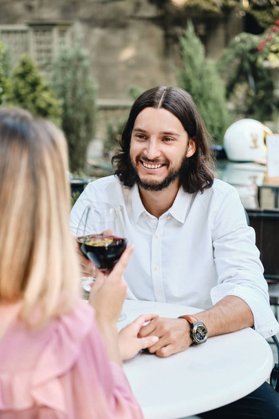 Joven atractiva morena barbuda hombre en camisa con copa de vino mirando alegremente a la novia en una cita romántica en la cafetería al aire libre
 - Foto, Imagen