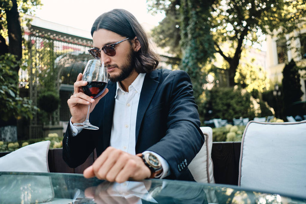 Joven morena confiada barbudo hombre cuidadosamente oler copa de vino tinto en el restaurante al aire libre
 - Foto, imagen