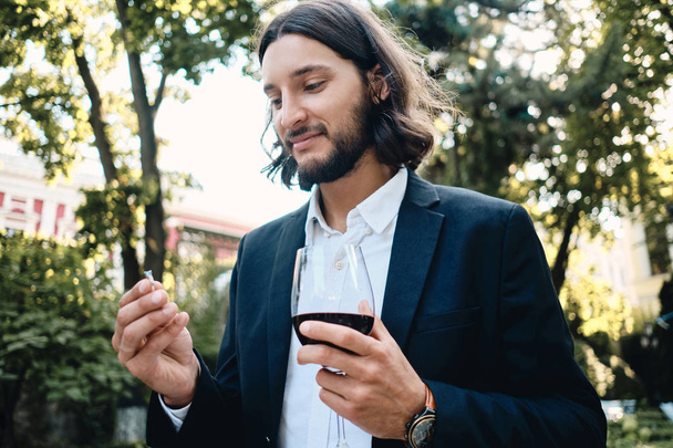 Young handsome brunette bearded man with glass of wine dreamily holding wedding ring in restaurant outdoor - Photo, Image