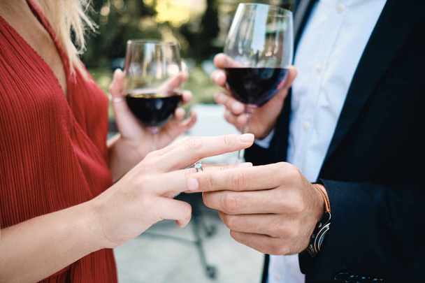 Close up jovem casal com copos de vinho colocando em anel de casamento no restaurante ao ar livre
 - Foto, Imagem