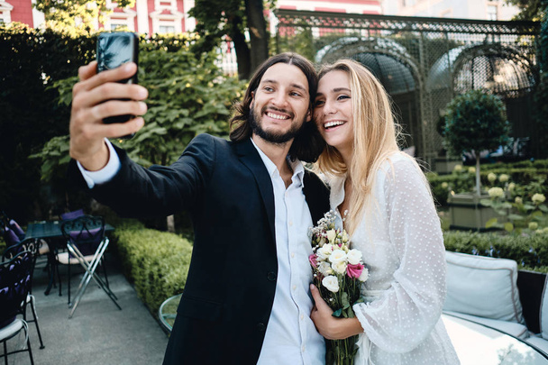 Młody wesoły narzeczony i narzeczona szczęśliwie biorąc selfie na telefon komórkowy razem w pięknym ogrodzie restauracji  - Zdjęcie, obraz