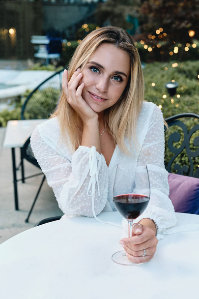 Jovem mulher loira bonita em vestido branco sonhadoramente olhando na câmera com copo de vinho no jardim do restaurante
 - Foto, Imagem