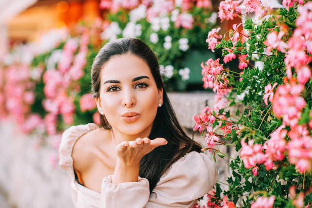 Primer plano retrato de una hermosa joven con el pelo largo y brillante, posando al aire libre en flores rosadas, enviando un beso
 - Foto, imagen