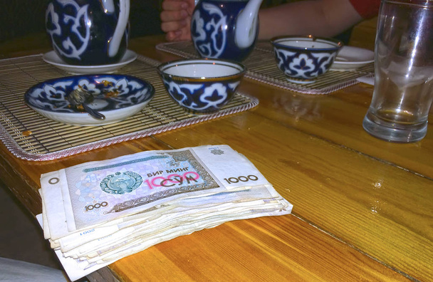 em uma mesa de madeira um conjunto de chá com um ornamento tradicional e um grande pacote de somas em dinheiro (a moeda do Uzbequistão), foco no dinheiro
 - Foto, Imagem
