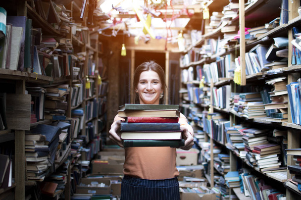 girl student holds a stack of books in the library, she searches for literature and offers to read, a woman prepares for study, knowledge is power, concept bookseller - Fotoğraf, Görsel