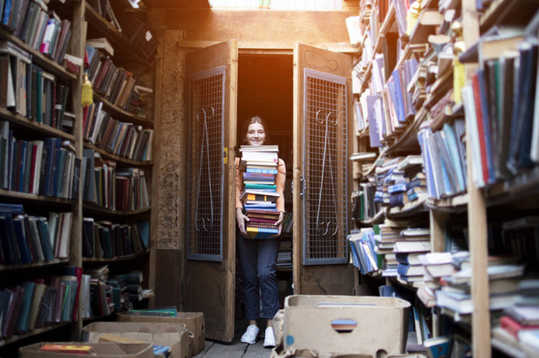 κορίτσι μαθητής κατέχει μια μεγάλη στοίβα βιβλίων και κουβαλάει πολλά λογοτεχνία στη βιβλιοθήκη, ετοιμάζεται για τη μελέτη, ο πωλητής των βιβλίων πήρε πολλά βιβλία με φόντο ένα βιβλιοπωλείο - Φωτογραφία, εικόνα
