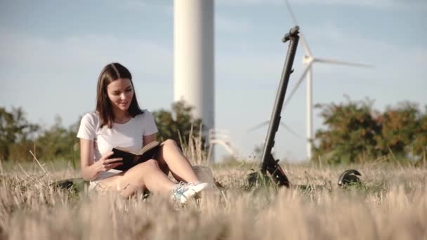 Een aantrekkelijk meisje zit naast een elektrische scooter tegen de achtergrond van windkracht stations en Leafs door middel van een boek. - Video