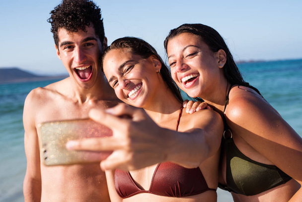 Две молодые женщины и молодой человек смеются и улыбаются в камеру мобильного телефона, когда они делают селфи на пляже
 - Фото, изображение
