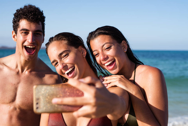 Due ragazze e un ragazzo ridono e sorridono alla fotocamera del telefono cellulare mentre si fanno un selfie sulla spiaggia
 - Foto, immagini