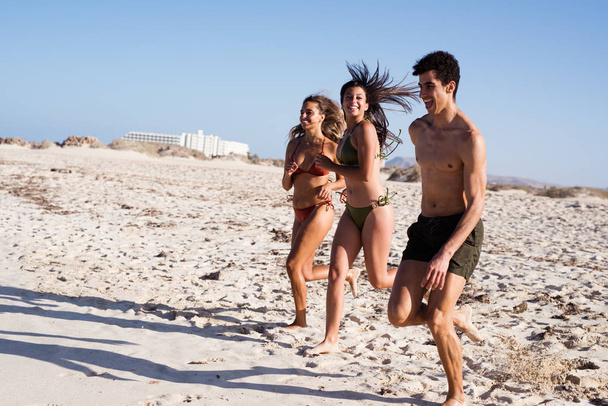 Two girls and a boy running on the beach as they laugh and smile in summer - Foto, Imagem