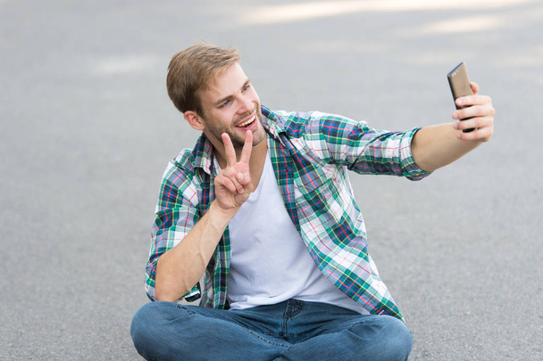 free time spending. summer fashion. happy man checkered shirt. male fashion. student relax use phone. macho man make selfie. street style. man sit on ground. carefree student. he love selfie - Фото, изображение