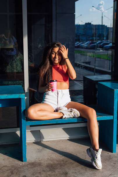 Stylish happy young brunette woman wearing white shorts and sneakers holding pink cup of coffee to go. - Photo, Image