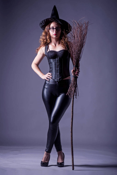 Sexy woman in leather pants and corset posing with a broom against a dark background. Witch in erotic clothing. - Zdjęcie, obraz