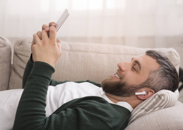 Cheerful man watching movies on digital tablet with wireless earphones - Foto, imagen