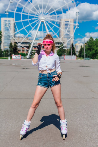 Portrait of an emotional girl in a pink cap visor and protective gloves for rollerblades and skateboarding riding on rollerblades on the road. - Фото, изображение