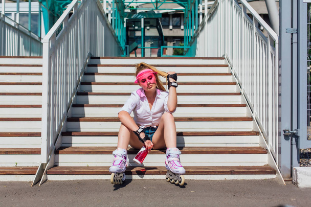 Portrait of an emotional girl in a pink cap visor wearing protective gloves and rollerblades sitting on stairs holding a bottle of water in hands - Фото, изображение