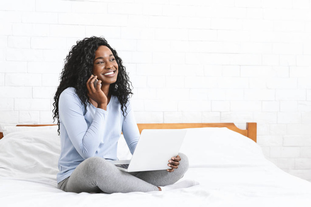 Woman sitting on bed in front of laptop, using cellphone - Photo, Image
