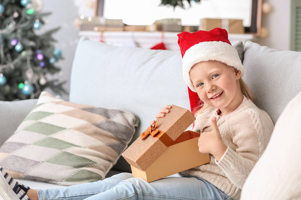 Χαριτωμένο μικρό κορίτσι με χριστουγεννιάτικο δώρο δείχνοντας αντίχειρας στο σπίτι - Φωτογραφία, εικόνα