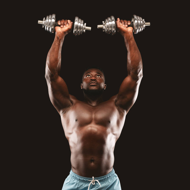 Heißer Bodybuilder hebt Hanteln vor schwarzem Hintergrund in die Höhe - Foto, Bild