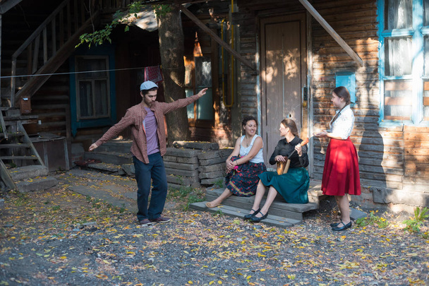 Un homme dansant la danse russe - des femmes assises dans les escaliers près de la maison de campagne et parlant entre elles - une femme jouant de la balalaika
 - Photo, image