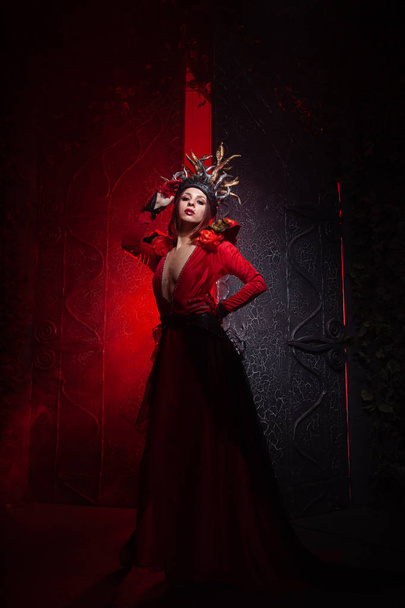 όμορφη γυναίκα μάγισσα της μόδας με κέρατα με τη μορφή ριζών δέντρων σε ένα μακρύ πολυτελές φόρεμα στο φόντο μιας τεράστιας πύλης με κόκκινο καπνό. Απόκριες ιδέα - Φωτογραφία, εικόνα
