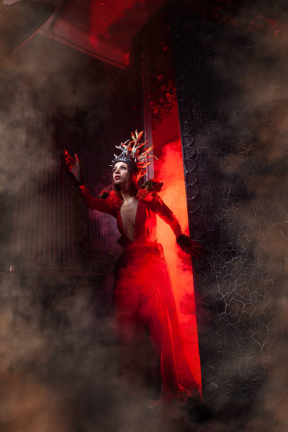piękna dziewczyna czarownica mody z rogami w postaci korzeni drzew w długiej luksusowej sukni na tle ogromnej bramy z czerwonym dymem. Koncepcja Halloween - Zdjęcie, obraz