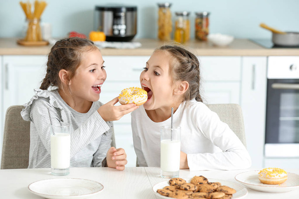 Portrait of cute twin girls eating donuts with milk in kitchen - Fotoğraf, Görsel