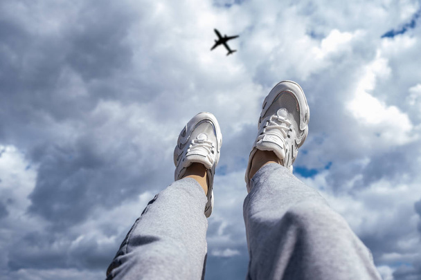 Nohy v bílých teniskách na pozadí oblačné oblohy a létající letadlo. Koncepce svobody. - Fotografie, Obrázek