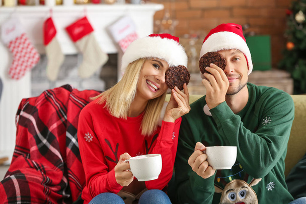 Ευτυχισμένο ζευγάρι στα καπέλα Σάντα πίνοντας ζεστή σοκολάτα και τρώγοντας μπισκότα στο σπίτι - Φωτογραφία, εικόνα