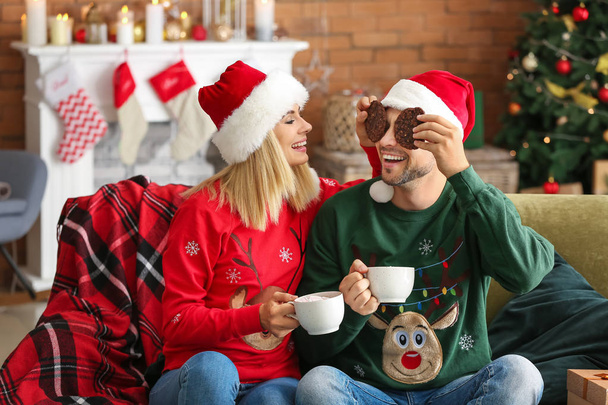 Happy couple in Santa hats drinking hot chocolate and eating cookies at home - Valokuva, kuva
