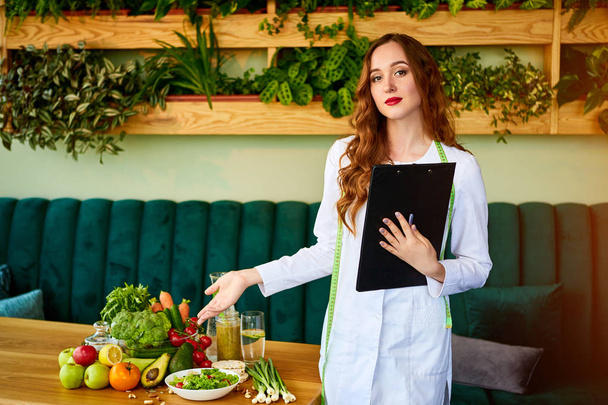Porträt einer jungen Ernährungsberaterin in medizinischem Gewand, die mit Klemmbrett und Maßband im Büro steht, im Hintergrund gesunde Ernährung. Gewichtsverlust und richtiges Ernährungskonzept - Foto, Bild