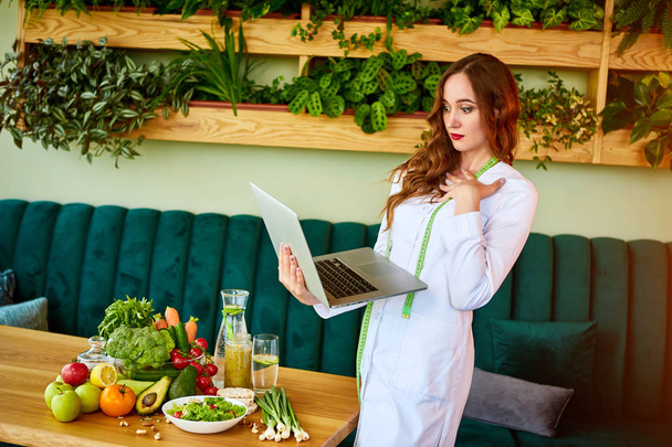 Kobieta dietetyka w mundurze medycznym z miara taśmy pracy z laptopem na planie diety stoi w pobliżu różnych zdrowych składników żywności w zielonym biurze na tle. Utrata masy ciała i prawo żywienia koncepcji - Zdjęcie, obraz