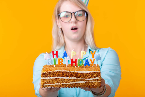 Portret van een grappig positief meisje met een papieren dop en een bril met een felicitatie cake in haar handen op een gele achtergrond. Concept en plezier en viering. Advertentieruimte. - Foto, afbeelding