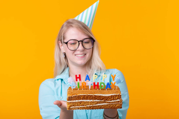 Holidays and party cocept - Positive young woman holding a happy birthday cake posing on a yellow background. - Photo, Image