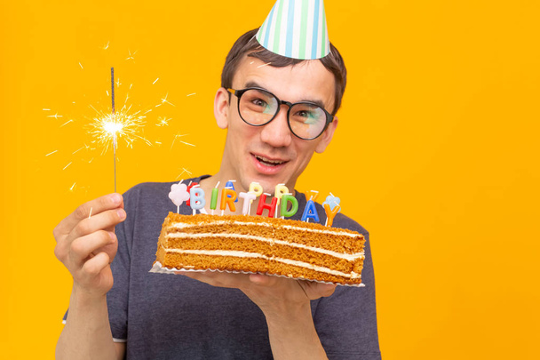 Positive funny young asian guy with a cap and a burning candle and a homemade cake in his hands posing on a yellow background. Anniversary and birthday concept. - Foto, Imagen
