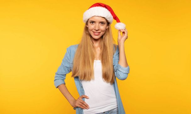 attractive young blonde woman in santa hat posing for Christmas holiday on yellow background - Photo, Image