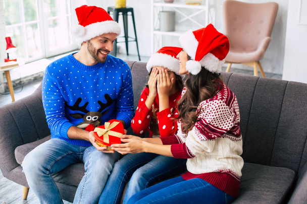 Mother and father in Santa hats giving daughter surprise gift for Christmas - Photo, Image