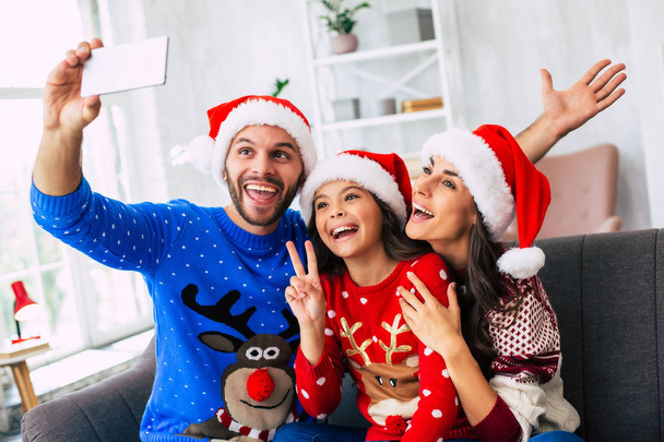 Family of mother, father and daughter in Santa hats posing for Christmas selfie on smartphone - Foto, afbeelding
