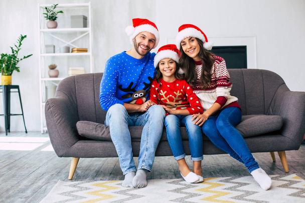 Family of mother, father and daughter in Santa hats and winter sweaters posing for Christmas holiday card of home sofa - 写真・画像