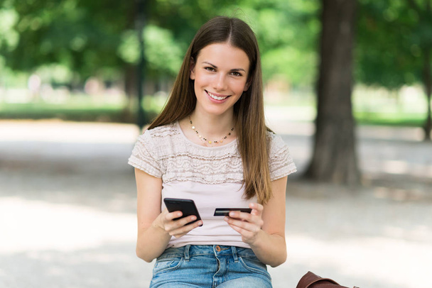 Kobieta robi zakupy online za pomocą jej karty kredytowej, siedząc w parku - Zdjęcie, obraz