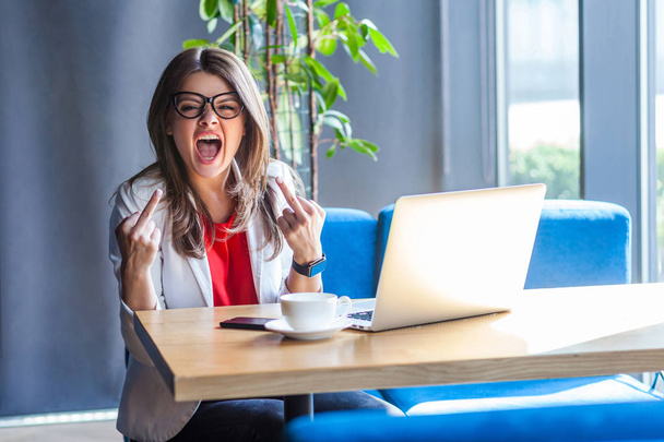 aggressive brunette young woman in glasses looking at camera and showing middle finger fuck signs and screaming with angry face while sitting at table with laptop in cafe, freelancing concept  - Foto, afbeelding